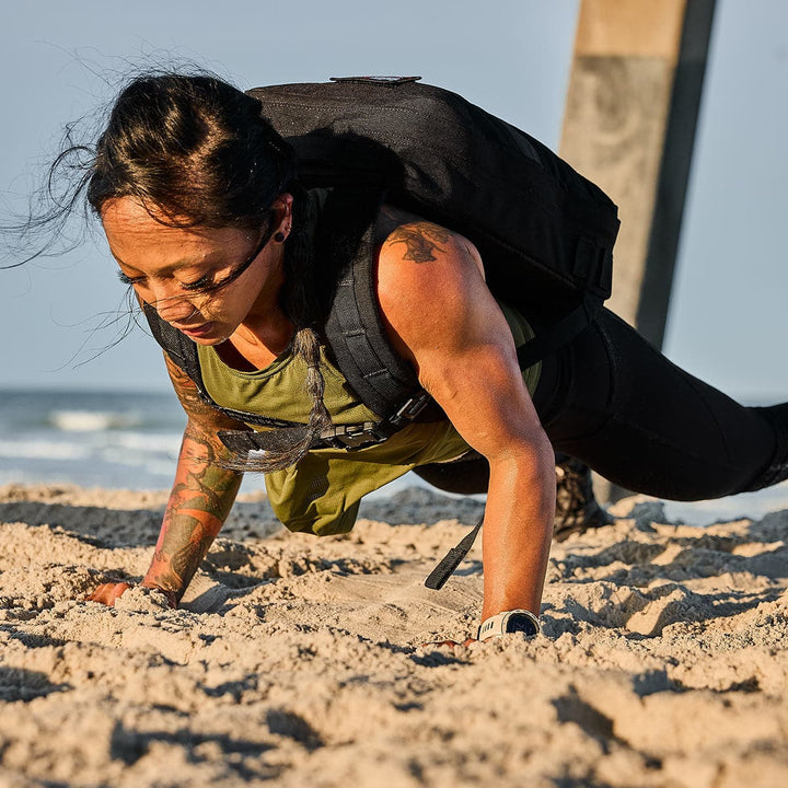 An individual is rucking on the beach, performing push-ups while wearing a weighted Rucker 4.0 backpack from GORUCK and a green tank top.