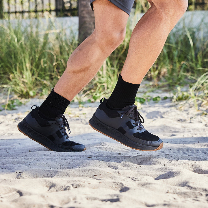 A person is running on a sandy beach in ultra-supportive GORUCK Rough Runner - Black + Gum shoes, featuring a Gradient Density™ EVA midsole, paired with black socks and gray shorts.