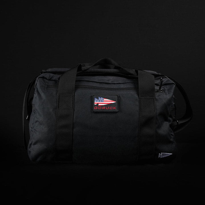 A black Kit Bag - Waxed Canvas by GORUCK, featuring handles and an American flag patch on the front, evocative of a Special Forces Deployment Bag, set against a dark background.