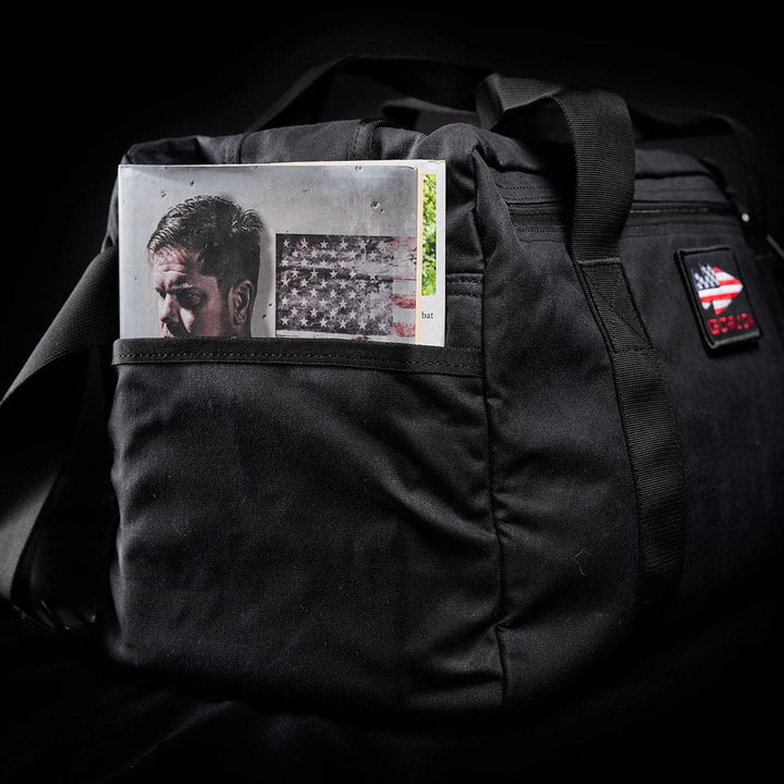 A sleek black Kit Bag - Waxed Canvas by GORUCK proudly displays a magazine with a man and flag emerging from the front pocket.