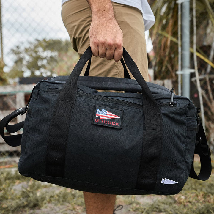 A person stands outdoors on a grassy area, holding a GORUCK Kit Bag - Waxed Canvas with an American flag patch.