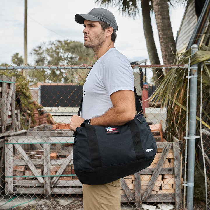 A man wearing a gray cap stands by a chain-link fence outdoors, carrying a black duffel that resembles the GORUCK Kit Bag - Waxed Canvas.