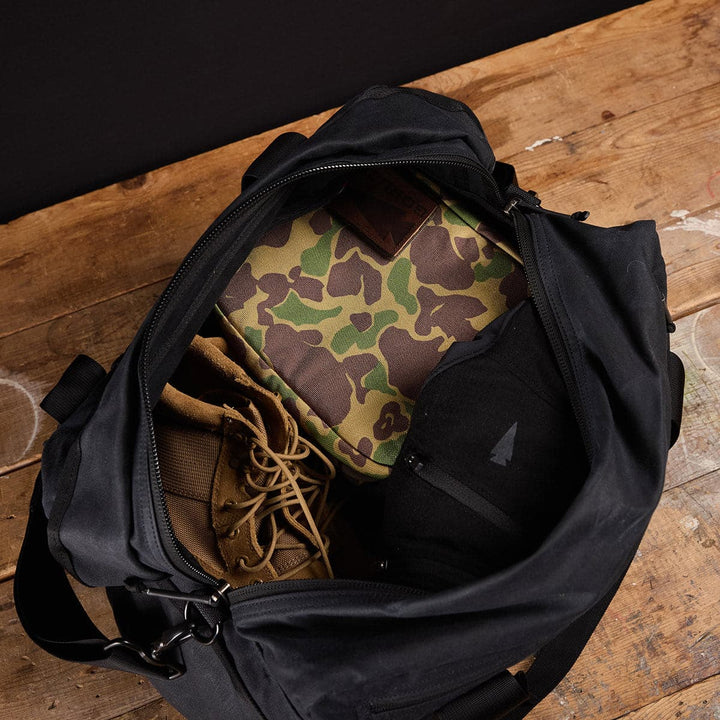 An open Kit Bag - Waxed Canvas by GORUCK reveals camouflage and black clothing, alongside sturdy brown boots, all set on a rustic wooden surface.