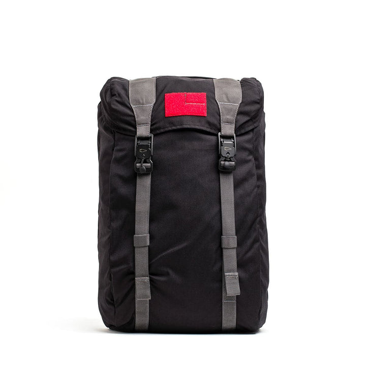 A sleek black GORUCK M23 - Cordura rucksack with gray straps and two buckles commands attention. Its minimalist design includes a striking red patch at the top, crafted from durable materials, standing out boldly against a white background.