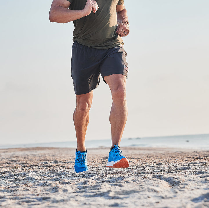 A man is jogging on a sandy beach, wearing a green shirt, dark shorts, and the Electric Blue Rough Runner by GORUCK. These shoes feature a Gradient Density™ EVA midsole for comfort and a versatile road-to-trail outsole perfect for any Rough Runner.