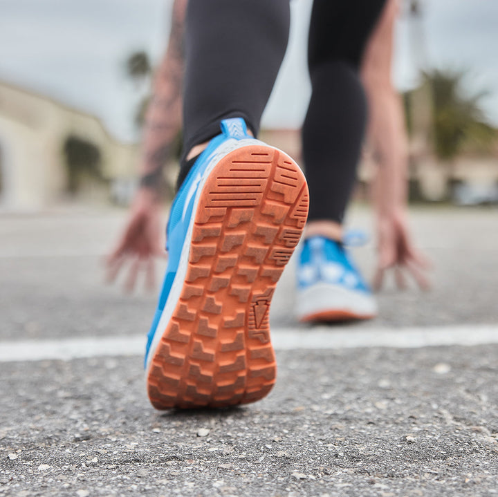 Close-up of a GORUCK Rough Runner - Electric Blue shoe on a track, preparing for a sprint, showcasing its GRADIENT DENSITY™ EVA MIDSOLE for optimal cushioning.