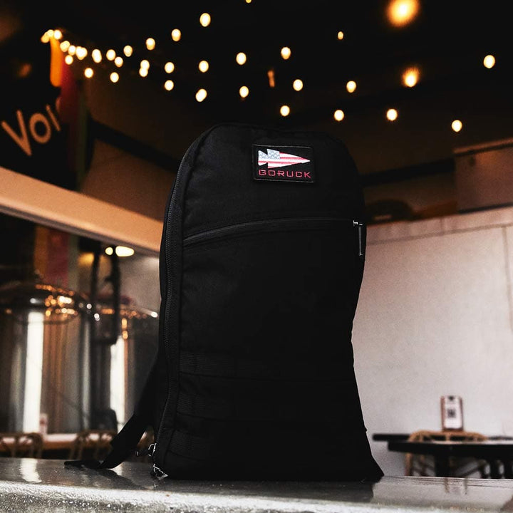 A sleek black backpack, the GORUCK Bullet Ruck Laptop - 16L, sits on the counter amid twinkling string lights. It boasts a bombproof laptop compartment and strong lumbar support, merging style with practicality for any adventure.