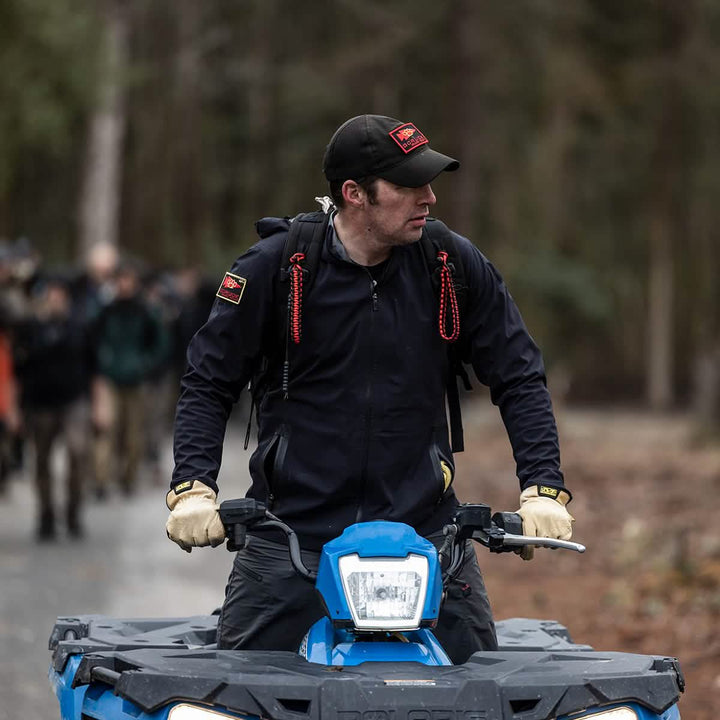 A person clad in a black cap and jacket rides a blue ATV through a forest, wearing Men’s Challenge Pants made of Lightweight ToughDry® fabric by GORUCK, known for their abrasion resistance. They gaze sideways as, in the blurry background, a group dressed in outdoor gear featuring articulated knees is walking.