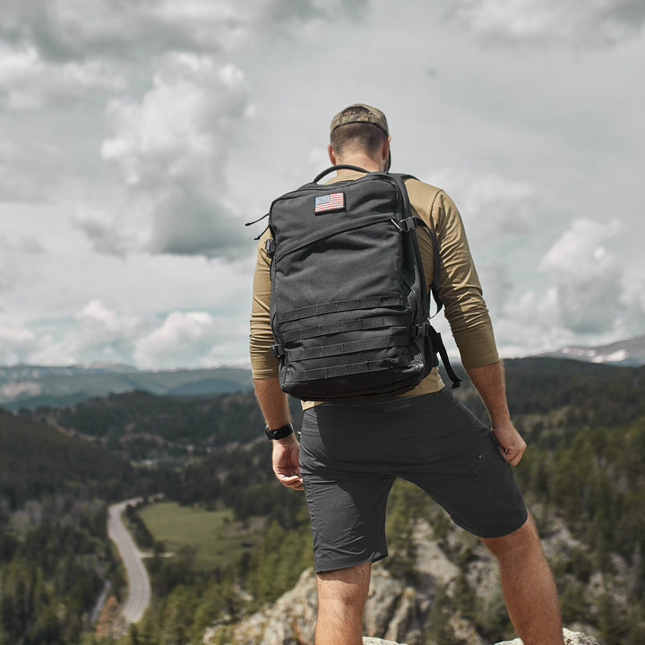 A person wearing GORUCK Men’s Challenge Shorts - Lightweight ToughDry® stands on a rocky ledge, their cargo pockets filled with essentials, overlooking a scenic landscape of forests and distant mountains under a cloudy sky.