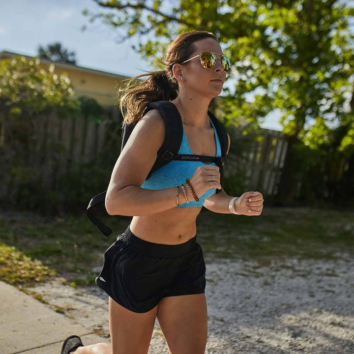 On a sunny day, someone jogs outdoors wearing sunglasses, a blue sports top, and black shorts. They look focused with the GORUCK Ruck Plate Carrier 3.0 on their back, featuring ergonomic lumbar support. The durable 1000D CORDURA material of the bag ensures comfort amidst a backdrop of green trees and a wooden fence.