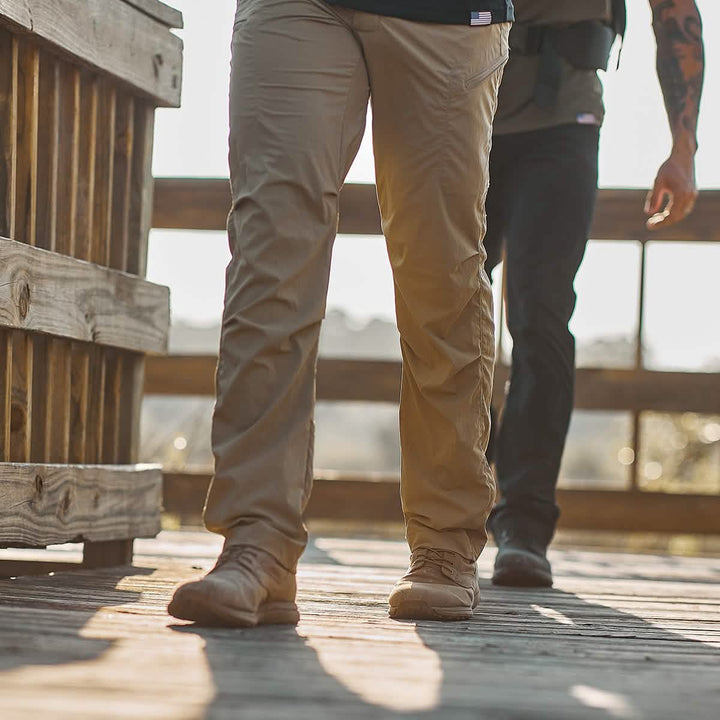 Two individuals walk on a wooden deck. The focus is on their legs and feet, clad in GORUCK’s Men’s Challenge Pants made from Lightweight ToughDry® material—one pair in khaki paired with boots, the other in black accompanied by dark shoes. Sunlight casts shadows on the deck, suggesting a sunny day outdoors.