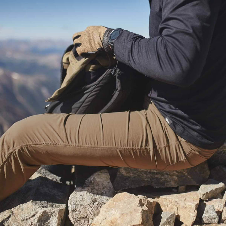 A person in a black jacket and Men’s Simple Pants from GORUCK, made with Lightweight ToughDry® material, sits on a rocky surface holding a backpack. They are wearing gloves and a wristwatch. The background features a mountainous landscape under a clear blue sky.