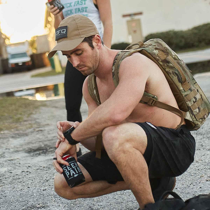 A man wearing a GORUCK Performance TAC Hat - TOUGHDRY and shorts kneels outdoors with a ToughDry® fabric rucksack, checking the time on his smartwatch while holding a can.