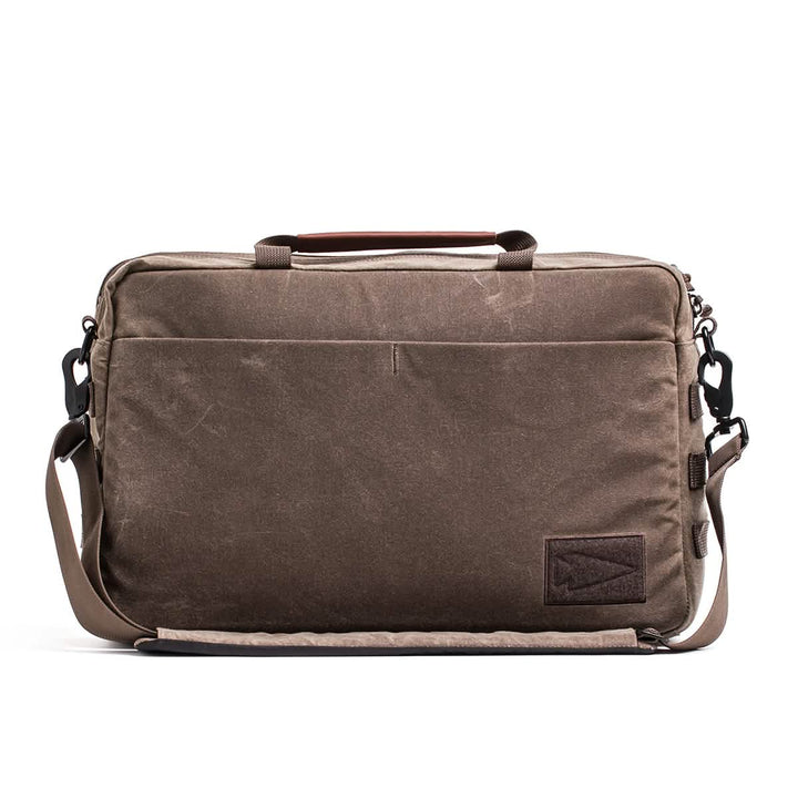 The GORUCK Shoulder Bag - Waxed Canvas is displayed in brown, featuring a professional design with a front pocket and adjustable strap. It includes a leather handle, side metal clasps, and a distinctive small leather patch with triangular stitching on the lower front corner, perfect for those looking for both style and efficiency in their daily carry.