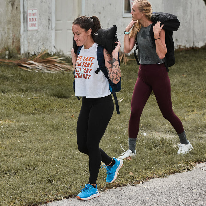 Two women, reminiscent of the GORUCK Rough Runner - Electric Blue, stride along the road with heavy bags on their shoulders. Their steps are confident, thanks to the versatile road-to-trail outsole that ensures each footfall is as stable off-road as it is on solid pavement.