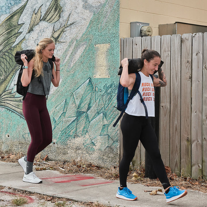 Two women, exemplifying the essence of GORUCK's Rough Runner - Electric Blue, stride confidently with heavy bags on their shoulders. As they walk past a vibrant mural, their shoes are equipped with a versatile road-to-trail outsole that ensures every step is seamless across different terrains.