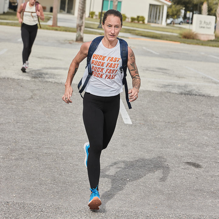 A person sprints through an empty parking lot, their "Ruck Fast" shirt catching the eye, while another figure lingers in the background. On their feet are GORUCK's Rough Runner shoes in Electric Blue, featuring a versatile road-to-trail outsole perfect for any terrain they encounter.