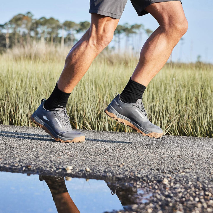 A person runs on a gravel path beside a reflective puddle, wearing GORUCK Mackall shoes in Forged Iron, Chiseled Stone, and Gum, complemented by gray outdoor shoes and black socks.