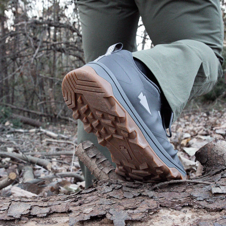 A person confidently steps over a tree log in the forest, showcasing their off-road prowess wearing GORUCK's Mackall shoes with Forged Iron, Chiseled Stone, and Gum.