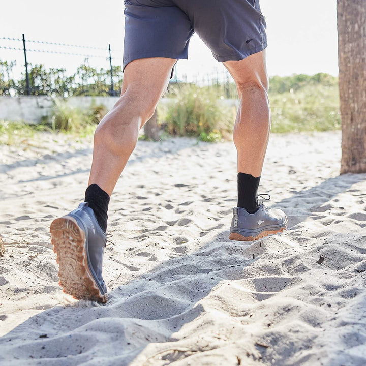 While jogging on sandy terrain near a palm tree, a person wears black socks, gray shorts, and GORUCK's Mackall in Forged Iron + Chiseled Stone + Gum, which have an all-terrain triple compound outsole.