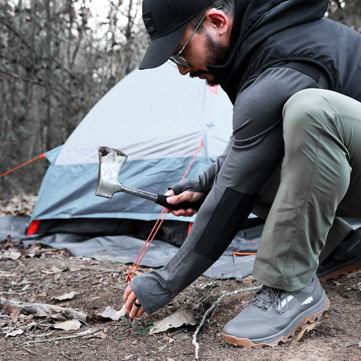 At a tree-filled campsite, someone in GORUCK Mackall shoes uses a hatchet to secure a tent stake in the ground.