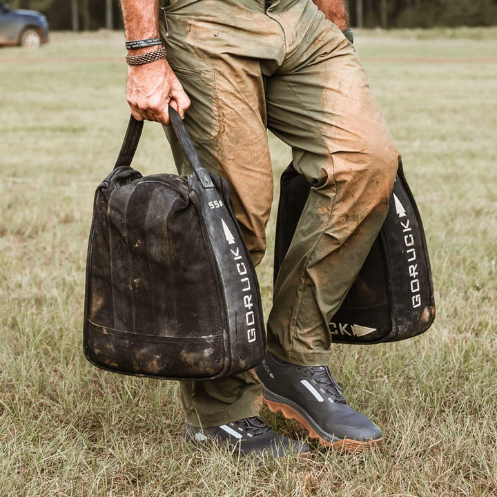 Outfitted like an off-road machine, a person carries muddy Mackall - Forged Iron + Chiseled Stone + Gum bags across the grass, their GORUCK pants and boots caked in mud.