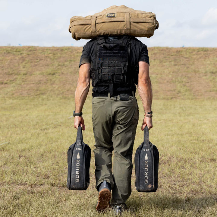 Wearing GORUCK's Mackall - Forged Iron + Chiseled Stone + Gum shoes, a person confidently strides across the grass with a large bag on their shoulders and two weights in hand, heading for a hill.