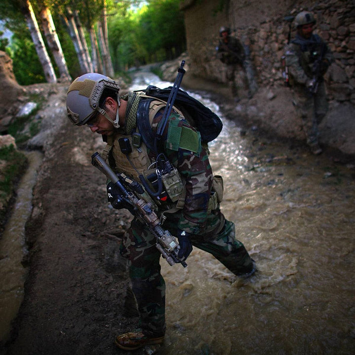 Wearing his GR1 USA from GORUCK securely strapped on, a soldier in a camouflage uniform crosses a stream with others trailing behind through a lush, tree-lined area.