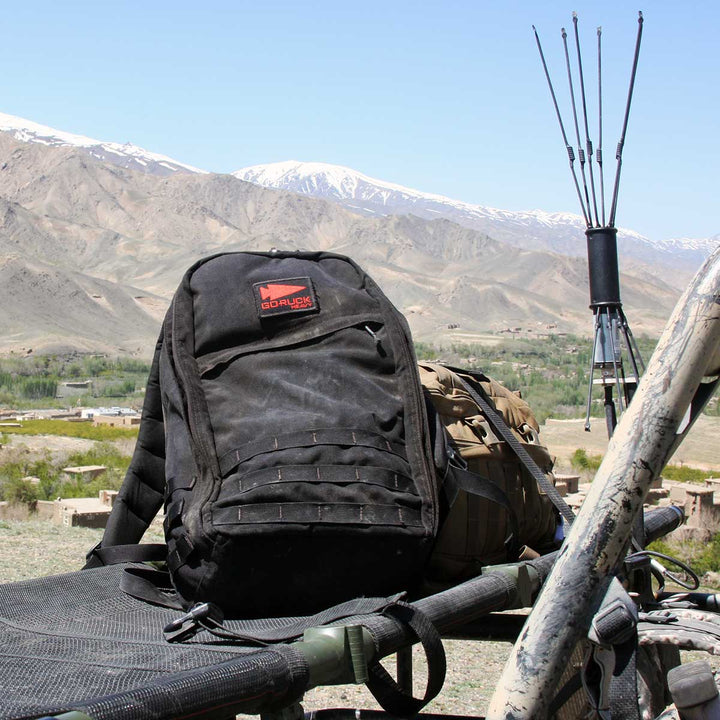 A black GR1 USA rucksack by GORUCK rests on a vehicle, overlooking a mountainous landscape with snow-capped peaks, crafted from durable 210D CORDURA.