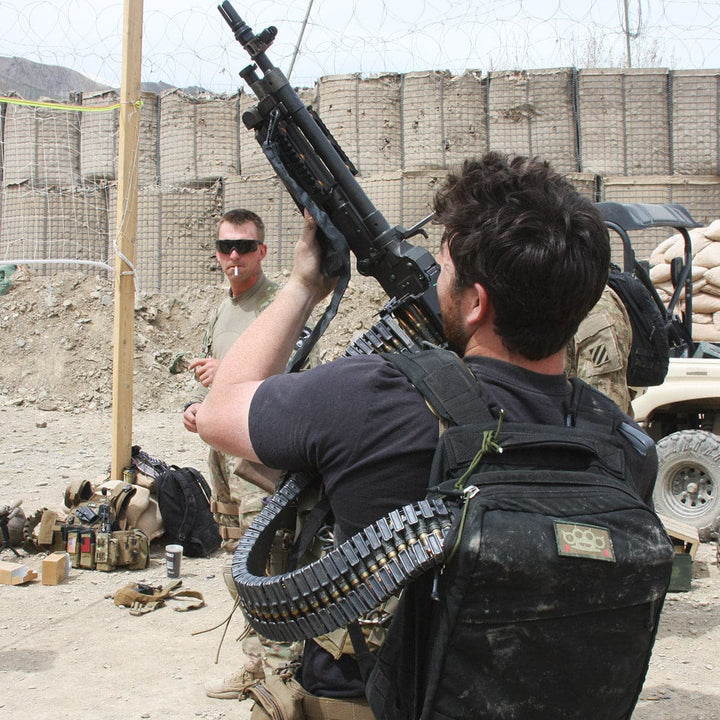 In an outdoor military setting, a man with a machine gun and ammunition belt stands ready, while another man lingers in the background. The rugged environment complements their durable GORUCK GR1 USA rucksacks, crafted from resilient 210D CORDURA fabric for toughness in demanding conditions.