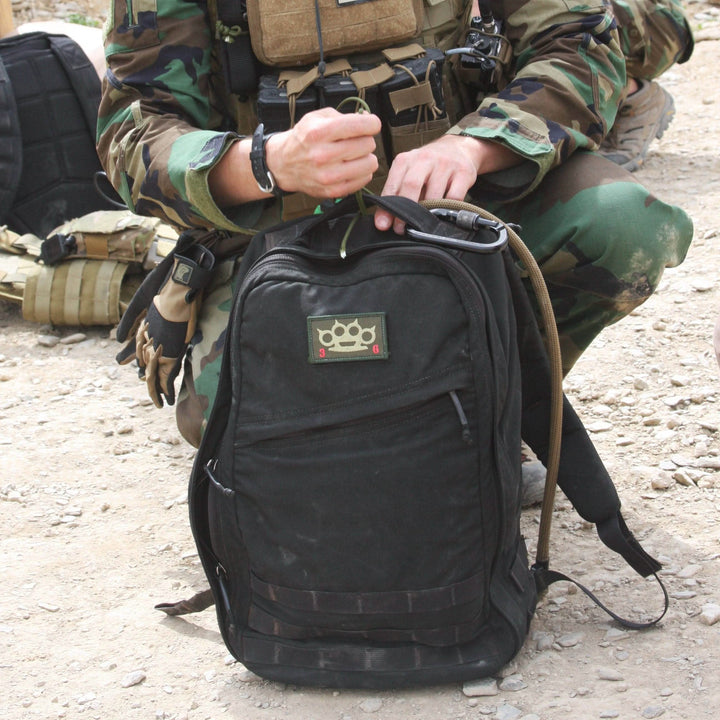 A person in camouflage gear kneels beside a black GR1 USA rucksack by GORUCK, made from durable 210D CORDURA, on a gravel surface.