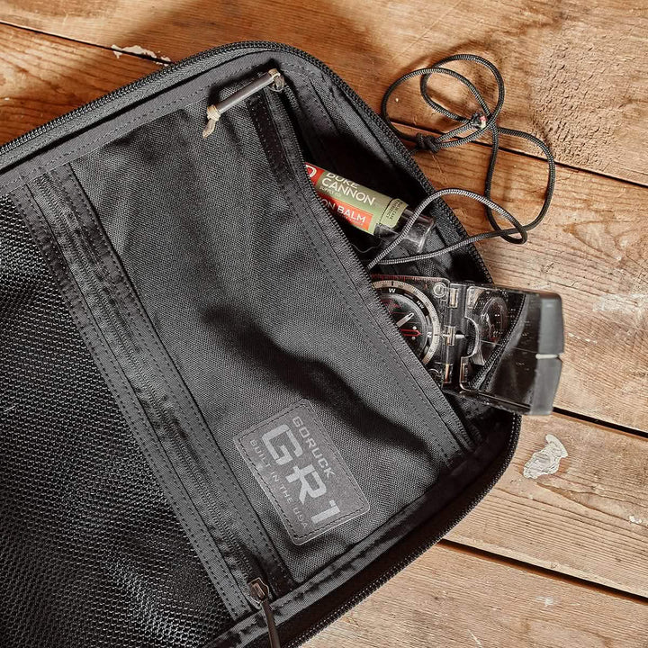 An open black GR1 USA CORDURA rucksack by GORUCK rests on a wooden surface, revealing a map, compass, and lip balm in its pocket.