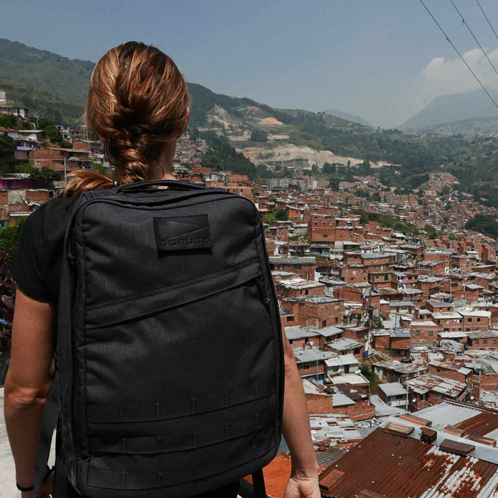 A person with a black GR1 USA rucksack by GORUCK gazes at a sprawling hillside city beneath a cloudy sky.