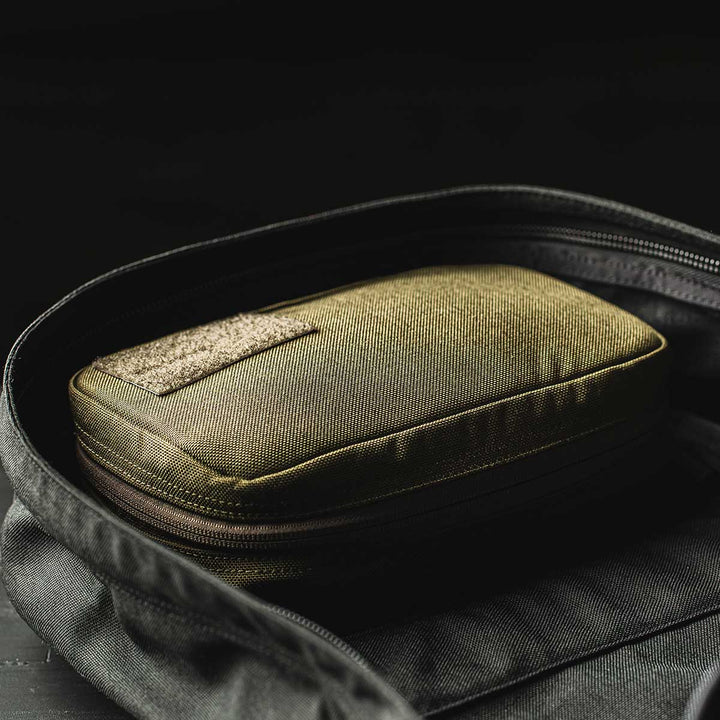 The GR1 Field Pocket by GORUCK, in a compact olive green design ideal for organizing electronics, peeks out from an open black bag against a dark background.