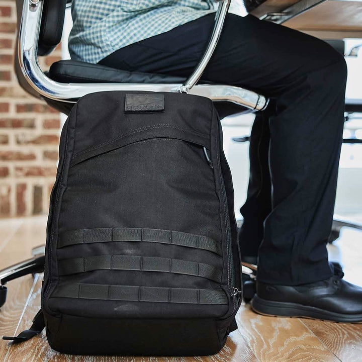 A sleek black GR1 USA rucksack by GORUCK, crafted from durable CORDURA fabric, rests on the floor beside a person at a desk in an office setting, showcasing its stylish and practical design.