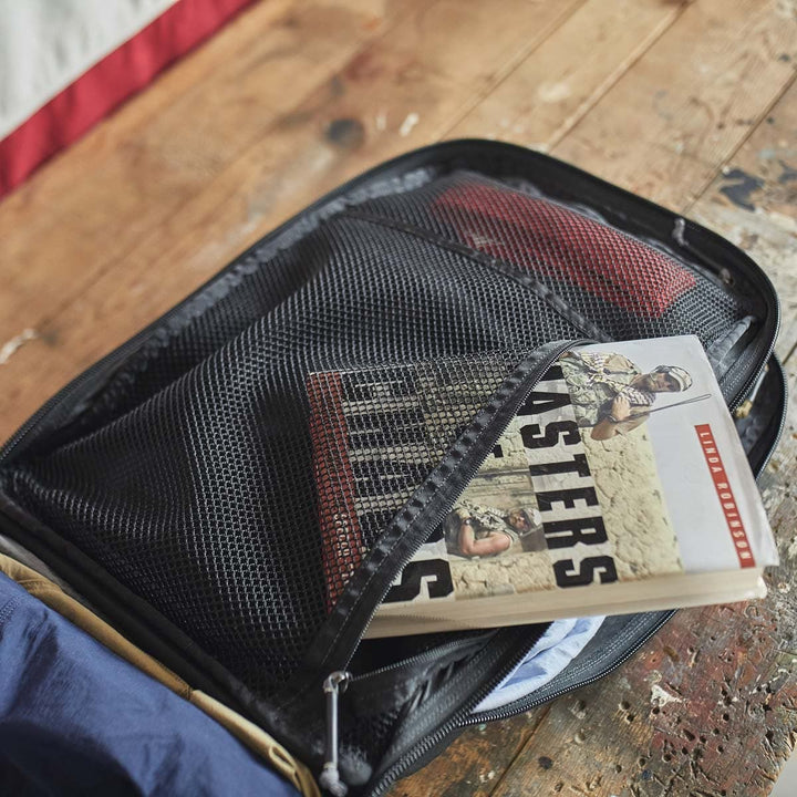 An open GR2 - USA by GORUCK rests on a wooden surface, its mesh pocket displaying a book titled "Masters of the Air." This stylish backpack is carry-on compliant, ideal for smooth and fashionable travels.