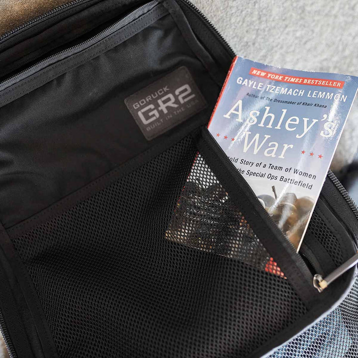 Placed inside the pocket of a GORUCK GR2 - USA black backpack, crafted from robust 210D CORDURA®, the book titled "Ashley's War" fits seamlessly with your travel essentials and is carry-on compliant.