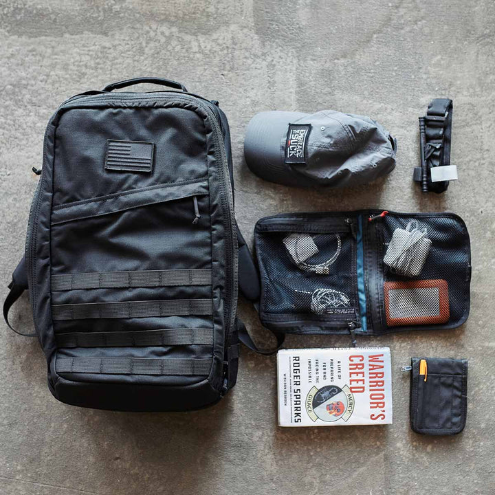 A black GR2 - USA backpack by GORUCK, crafted from resilient 210D CORDURA® fabric, is displayed on a concrete floor alongside a matching cap, book, and assorted gear—ideal for your next carry-on compliant adventure.