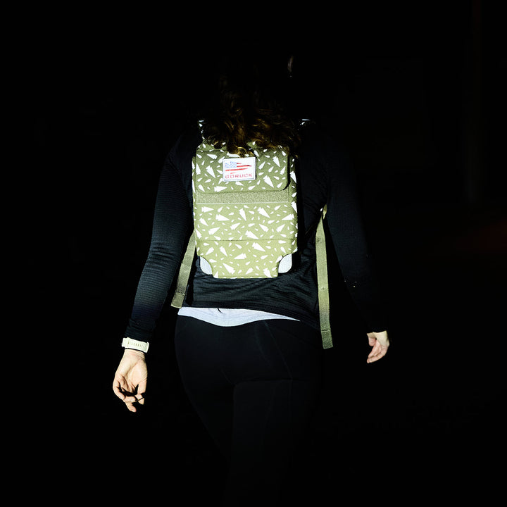 In the dim light, a person walks with a Ruck Plate Carrier 3.0 by GORUCK, featuring a green pattern with reflective elements and ergonomic lumbar support.