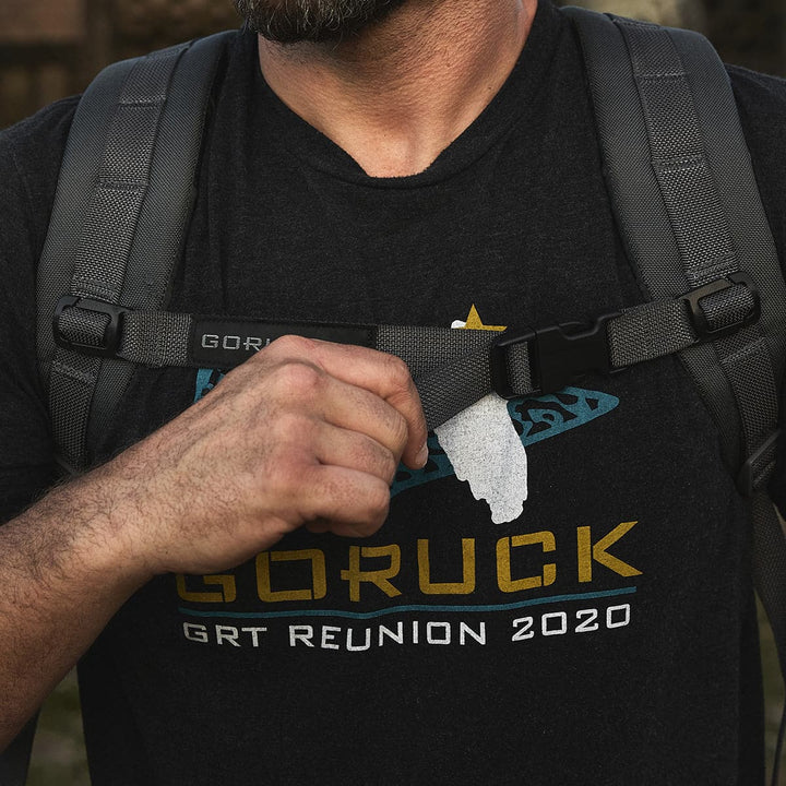 A man wearing a "Goruck GRT Reunion 2020" T-shirt adjusts the chest strap of his Ruck Plate Carrier 3.0 from GORUCK, highlighting its durable ballistic nylon construction and ergonomic lumbar support.