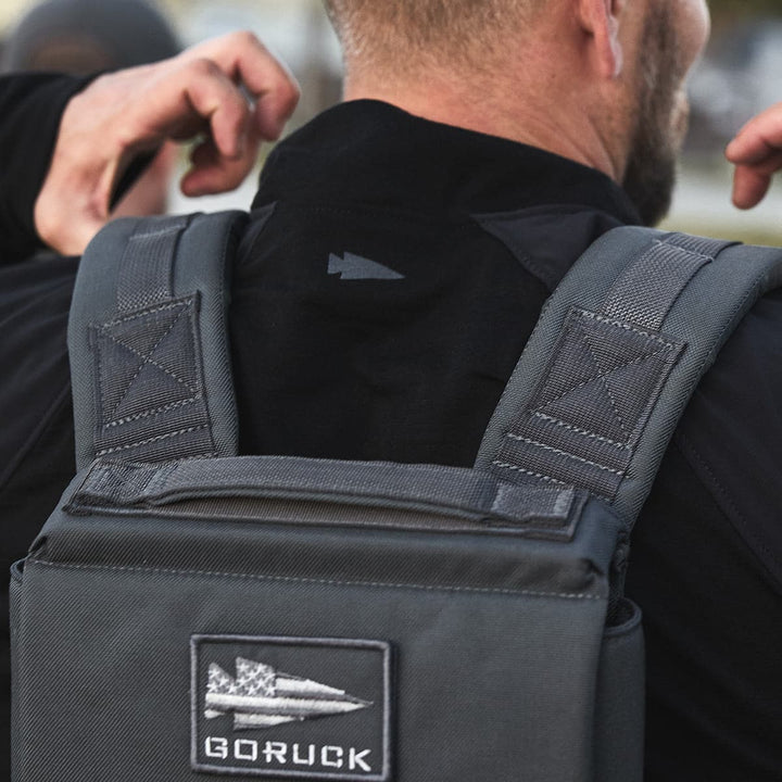 A man wearing the Ruck Plate Carrier 3.0 from GORUCK, crafted from durable ballistic nylon, adjusts the shoulder strap for comfort.