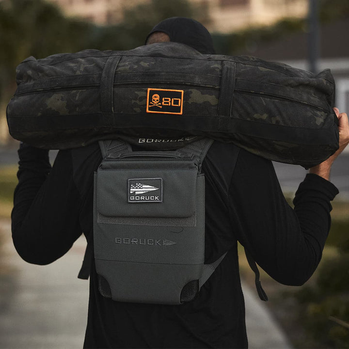 A person is carrying a heavy black sandbag on their shoulders, along with a GORUCK Ruck Plate Carrier 3.0 backpack, expertly designed for added comfort with ergonomic lumbar support.