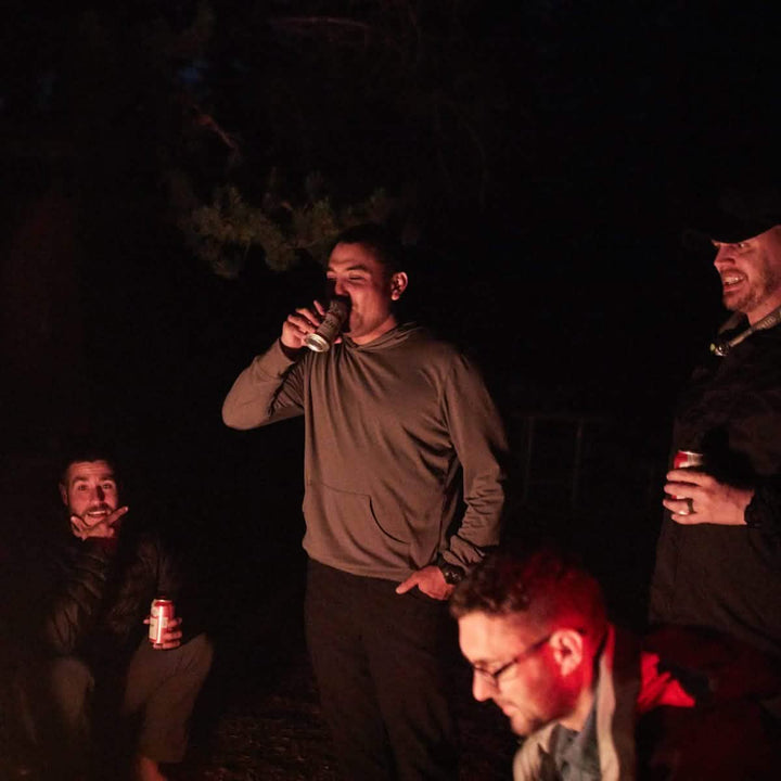 Four people are gathered around a campfire at night. One person, dressed in Men’s Simple Pants - Lightweight ToughDry® by GORUCK, is drinking from a can while others hold drinks, illuminated by the fire's glow. The scene is casual and relaxed, with trees visible in the background.