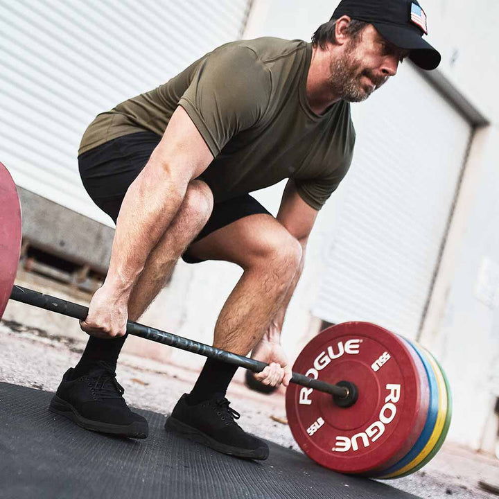 A man in a green shirt confidently steps into his workout shoes, GORUCK's Ballistic Trainers - Blackout + Black, and lifts a colorful barbell outdoors on a black mat. With 3X Support™ technology, he's well-equipped for functional fitness challenges.