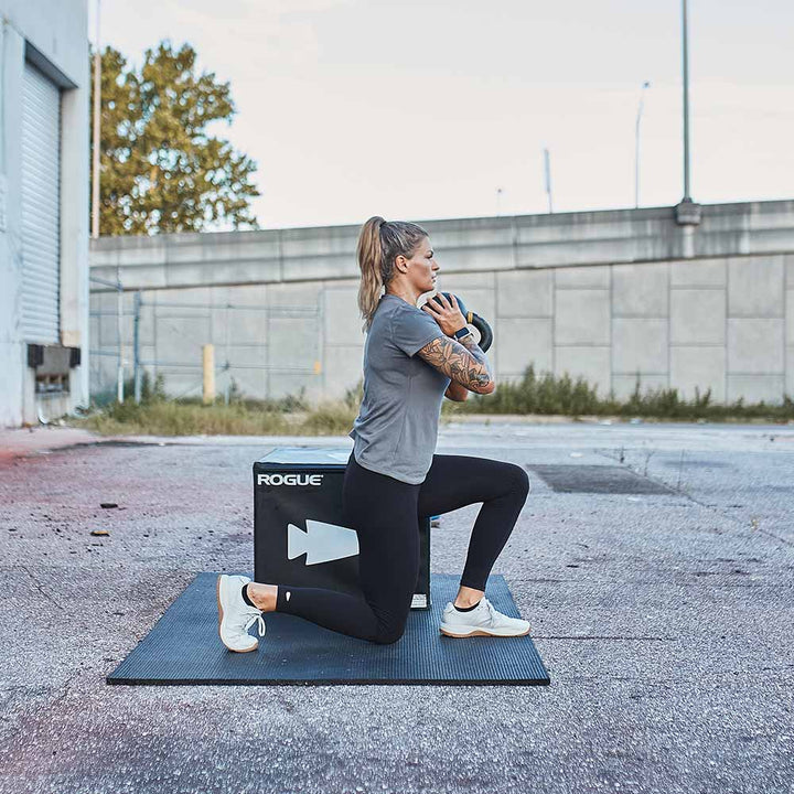 A person performs a kettlebell lunge on a mat outdoors, dressed in athletic clothing and wearing GORUCK Ballistic Trainers - Lunar Rock. These shoes feature 3X Support™ and CORDURA® Ballistic Nylon, providing stability and durability during the dynamic workout.