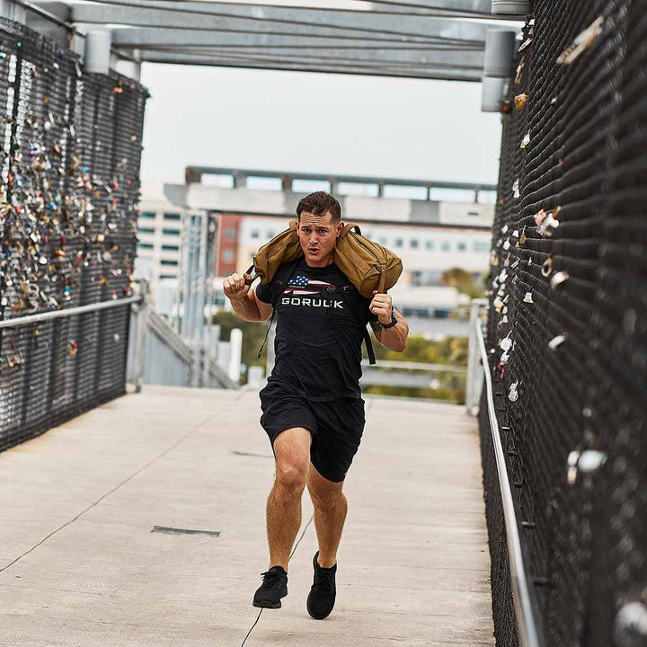 A man, embodying the essence of functional fitness, runs across a bridge with a sandbag over his shoulders, showcasing his determination in GORUCK's Ballistic Trainers - Blackout + Black workout shoes designed for optimal performance.