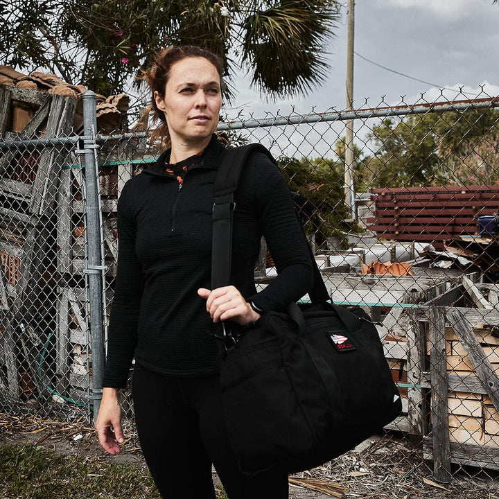 A woman stands outside holding a black GORUCK Kit Bag (Includes Shoulder Strap). She is wearing a black long-sleeve top and pants. The background includes a chain-link fence, wooden debris, and palm trees under a cloudy sky.