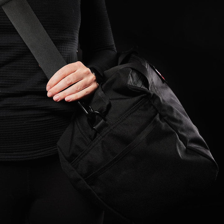 A person dressed in black holds a GORUCK Kit Bag (Includes Shoulder Strap) against a dark background. The bag, featuring a front zipper pocket and resembling an Aviator’s Kit Bag, is being securely held with one hand.