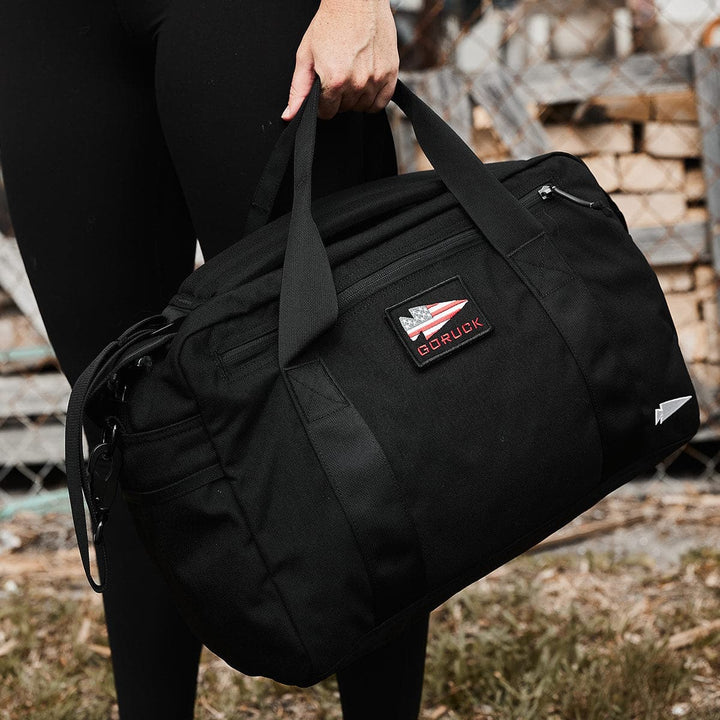 A person stands outdoors on grass, holding a GORUCK Kit Bag (Includes Shoulder Strap) that resembles deployment bags. The bag, featuring multiple handles, is positioned in front of a chain-link fence and stacked wooden planks. The individual is wearing black leggings.