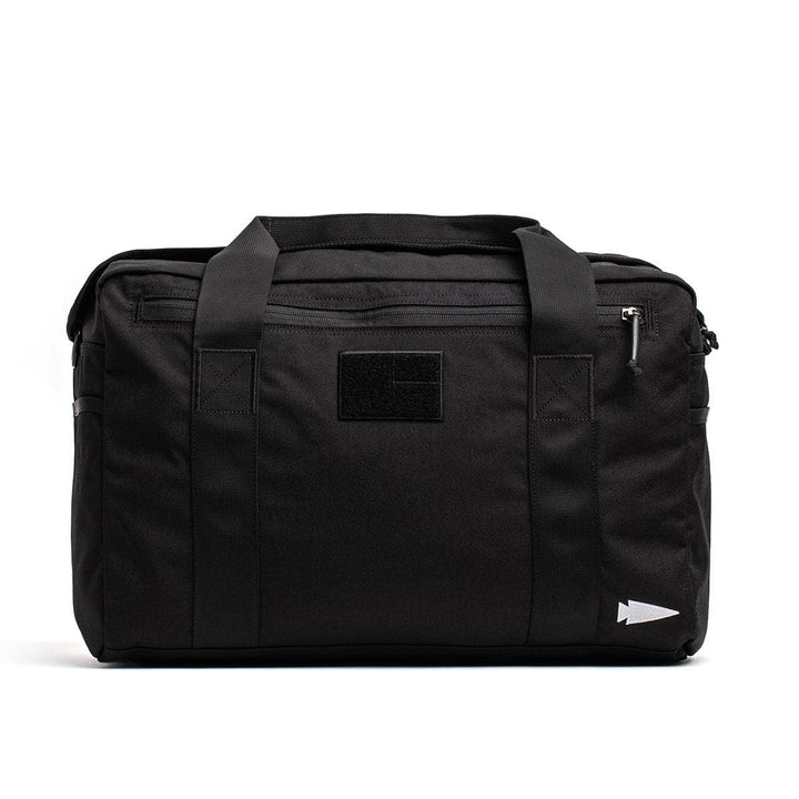 The Kit Bag by GORUCK, available in black and including a shoulder strap, is modeled after a Special Forces Flight Bag. It features sturdy handles and a front zipper pocket. The design is complemented by a small arrow logo positioned on the bottom right corner and a patch area centered at the top, all set against a plain white background.